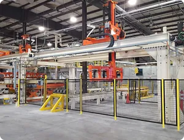 Industrial facility with automated machinery, including robotic arms and conveyor systems, enclosed by safety fencing and located in a spacious factory setting with overhead lighting.